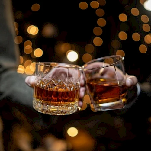 Two people clink whiskey glasses in a dim, festive setting with warm bokeh lights in the background.