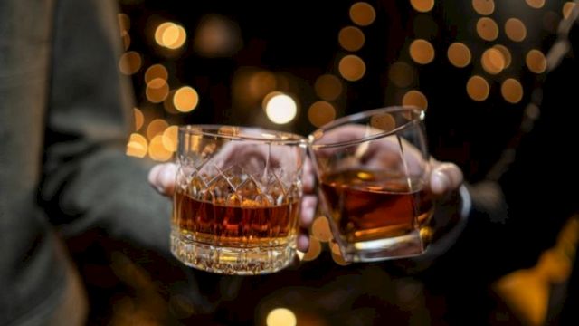 Two people clink whiskey glasses in a dim, festive setting with warm bokeh lights in the background.