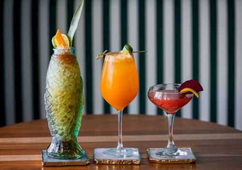 Three colorful tropical cocktails on a wooden bar, each in a tall or stemmed glass with fruit garnishes, against a striped background.