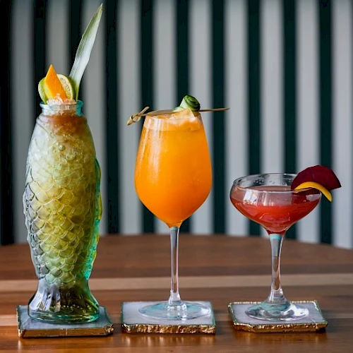 Three colorful tropical cocktails on a wooden bar, each in a tall or stemmed glass with fruit garnishes, against a striped background.