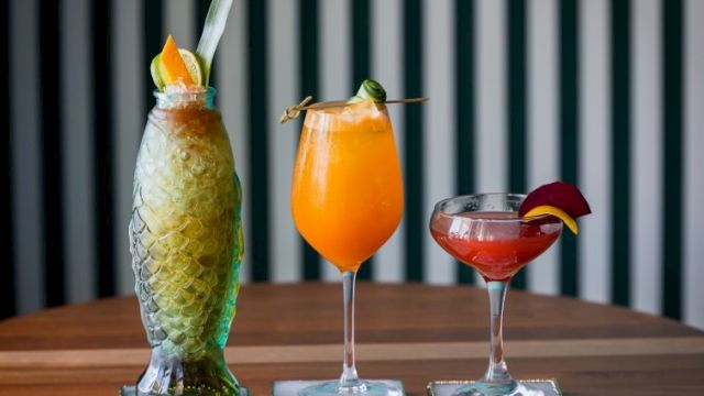 Three colorful tropical cocktails on a wooden bar, each in a tall or stemmed glass with fruit garnishes, against a striped background.