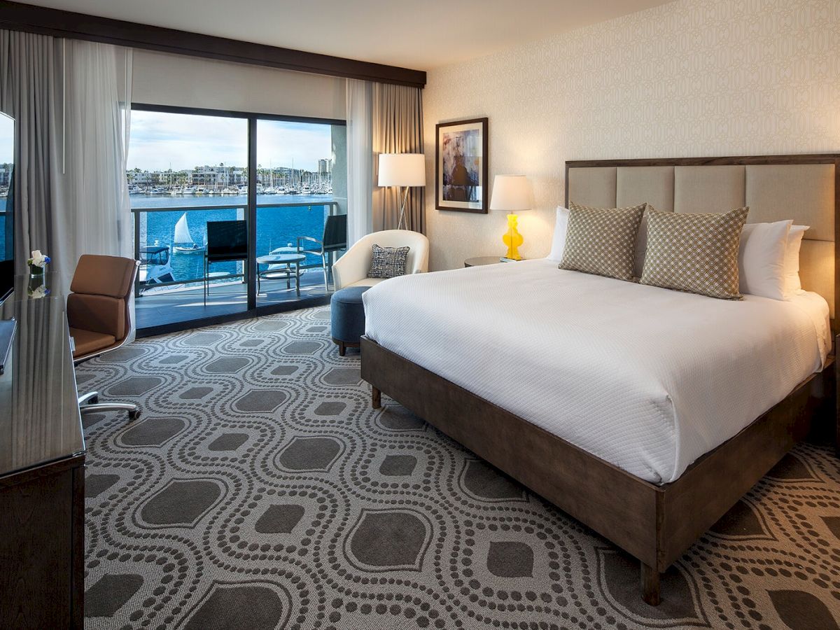 A modern hotel room with a large bed, two nightstands, a seating area, and a balcony overlooking water, featuring patterned carpet and soft lighting.