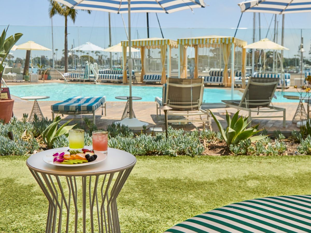 A sunny resort pool scene with striped chairs, umbrellas, and a small round table holding drinks and fruit on a grassy lawn. Ends with a period.