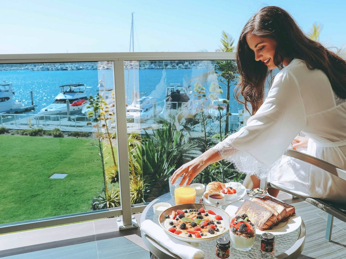 A woman enjoys a lavish breakfast on a balcony overlooking a marina, with pastries, fruit, yogurt, and a bright sea view, morning bliss.