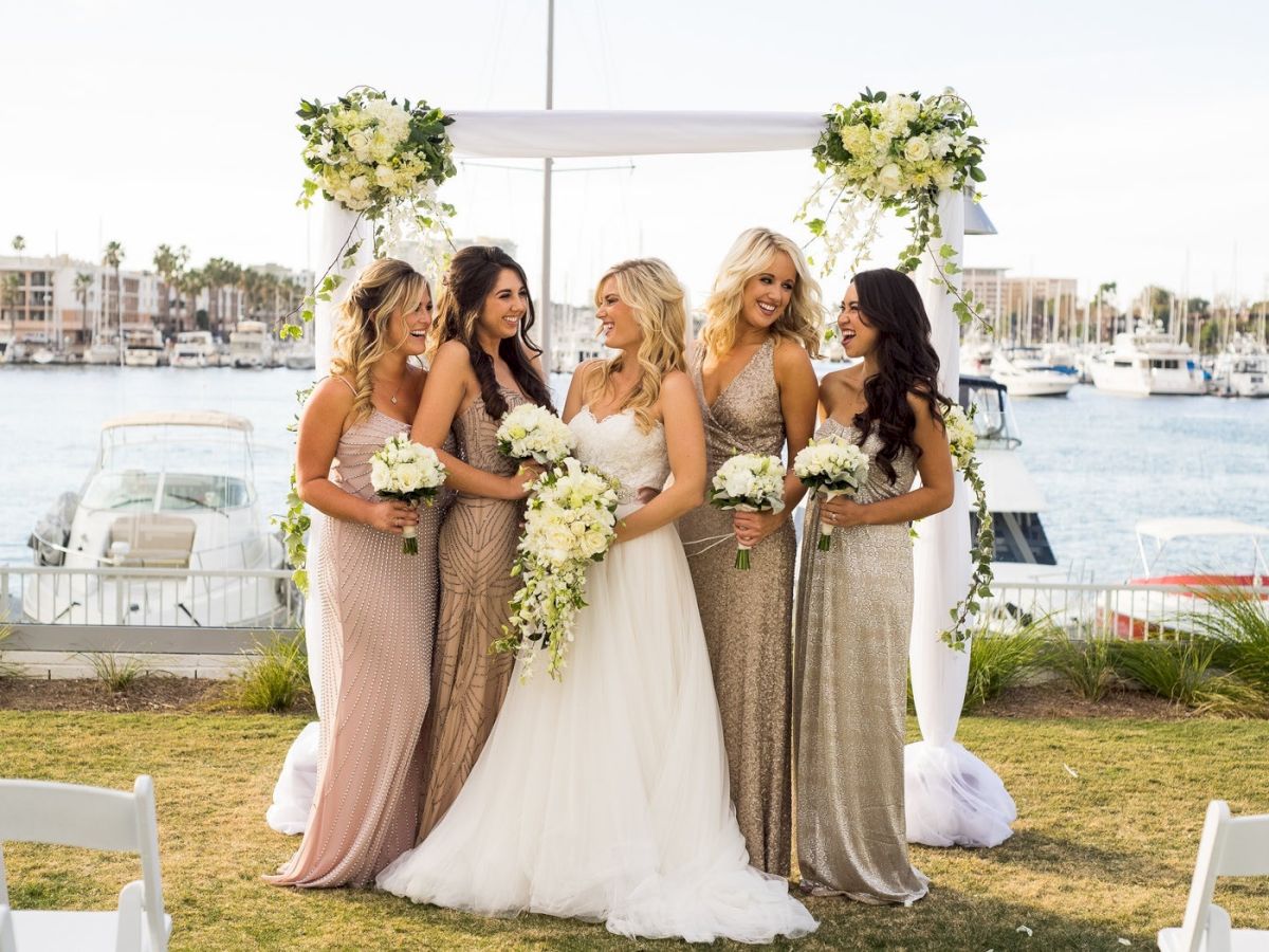 Six women at a seaside wedding: the bride in white gown flanked by bridesmaids in shimmering gowns, holding bouquets, with a harbor backdrop.