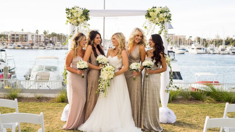 Six women at a seaside wedding: the bride in white gown flanked by bridesmaids in shimmering gowns, holding bouquets, with a harbor backdrop.