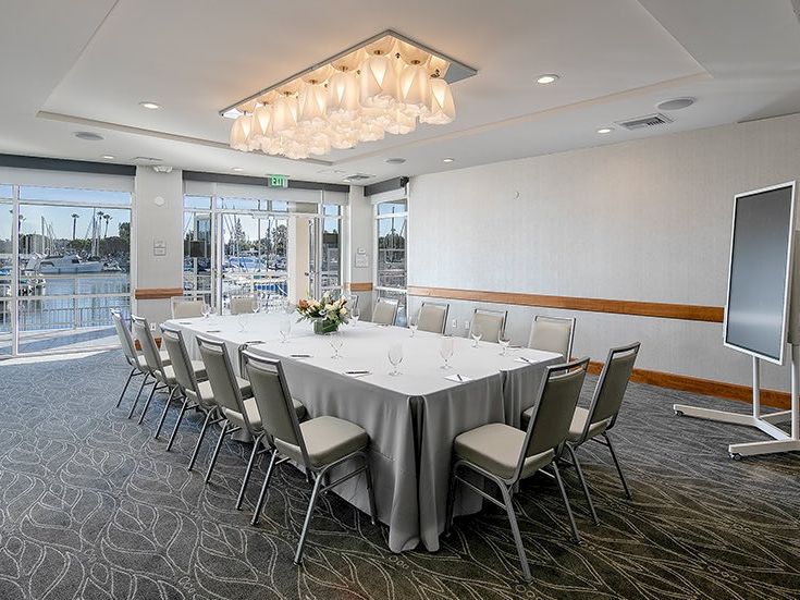 A formal meeting room set for a conference, with a long U-shaped table, grey chairs, a floral centerpiece, bright chandelier, and a view of the water outside.