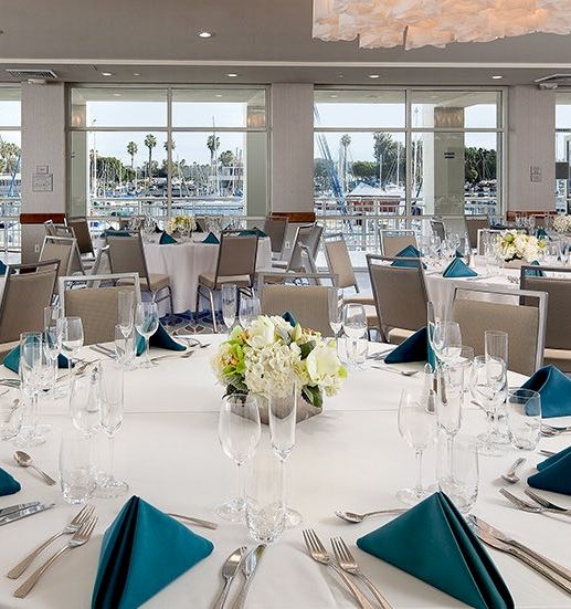 A bright banquet hall set for a formal event with round tables, white tablecloths, teal napkins, glassware, and a chic floral centerpiece.