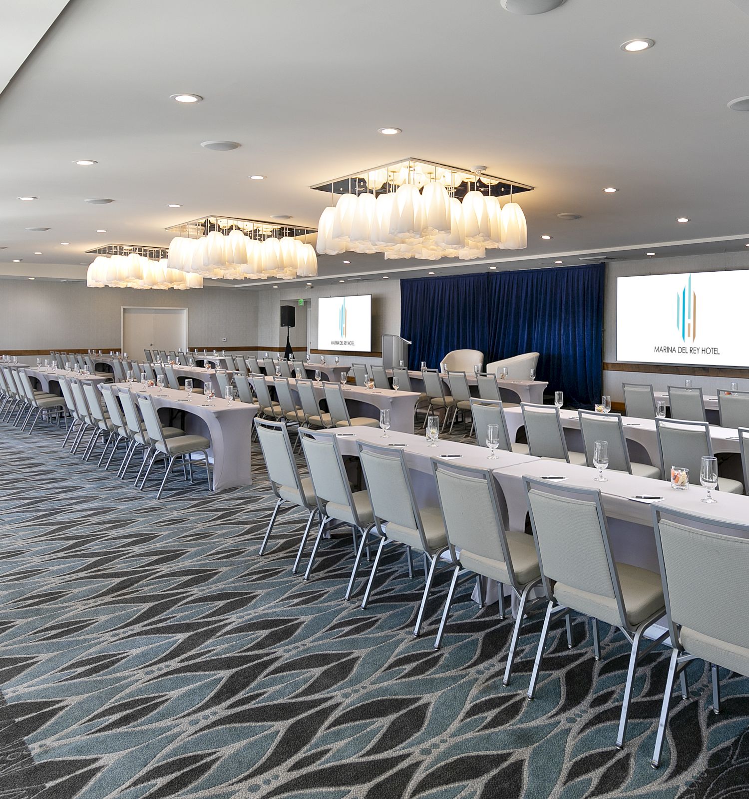 A spacious conference room set up with long rows of white-clothed tables and chairs, a stage with projector screens, and chandeliers overhead.
