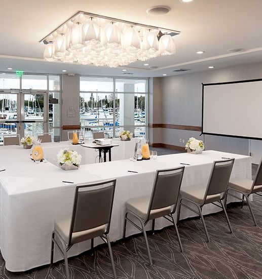 A conference room setup with a long U-shaped table, white tablecloths, flower centerpieces, a projector screen, and a bright, modern dining-style ambiance.