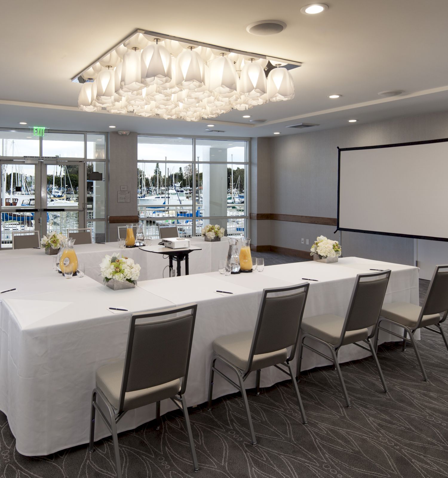 A conference room with a long rectangular table set for a meeting, white tablecloths, floral centerpieces, chairs, a projector screen, an easel, and large windows letting in natural light.
