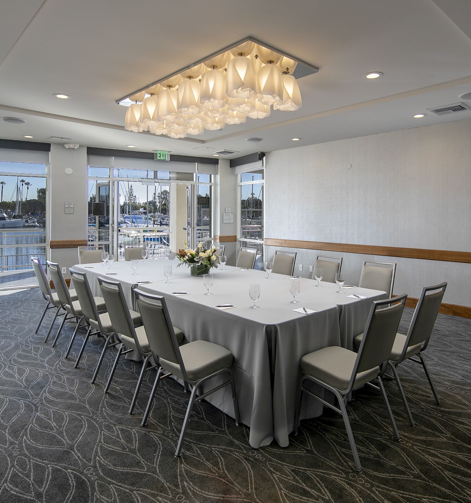 A modern conference room set for a meeting, with a long U-shaped table, gray chairs, a chandelier, and large windows overlooking a marina or water view, ending here.