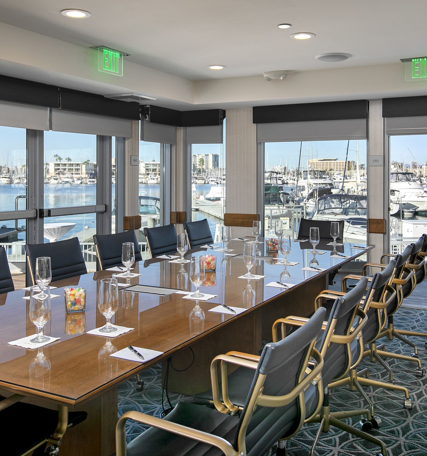 A conference room with a long wooden table, chairs, wine glasses, and place settings, overlooking a marina through large windows.
