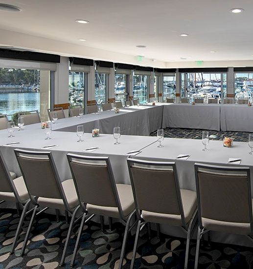 A bright, modern harbor-side conference room with a U-shaped table, grey chairs, large windows, and place settings for a formal meeting.