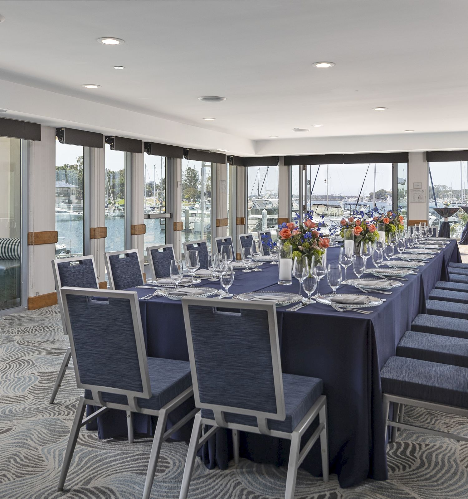 A long banquet table with dark blue tablecloths and neatly arranged place settings sits in a bright, glass-walled event room overlooking the water, ready for a formal gathering.