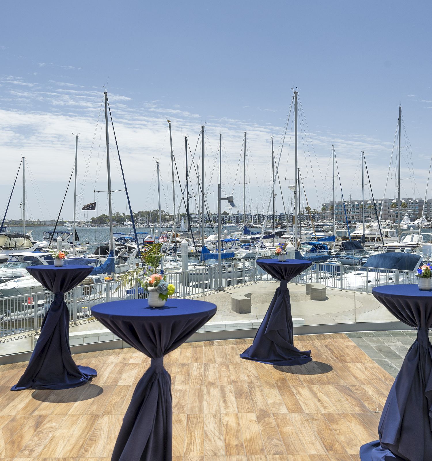 A marina with numerous sailboats docked, and tall cocktail tables draped in dark blue cloth by a wooden deck.