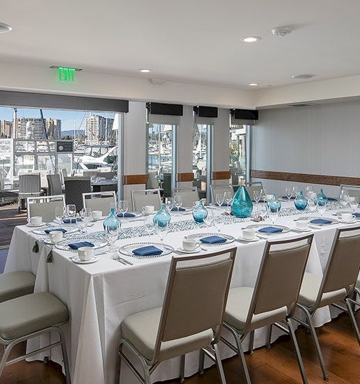 A formal meeting or banquet setup with a long, U-shaped table, white linens, place settings, turquoise glassware, and a marina view outside.
