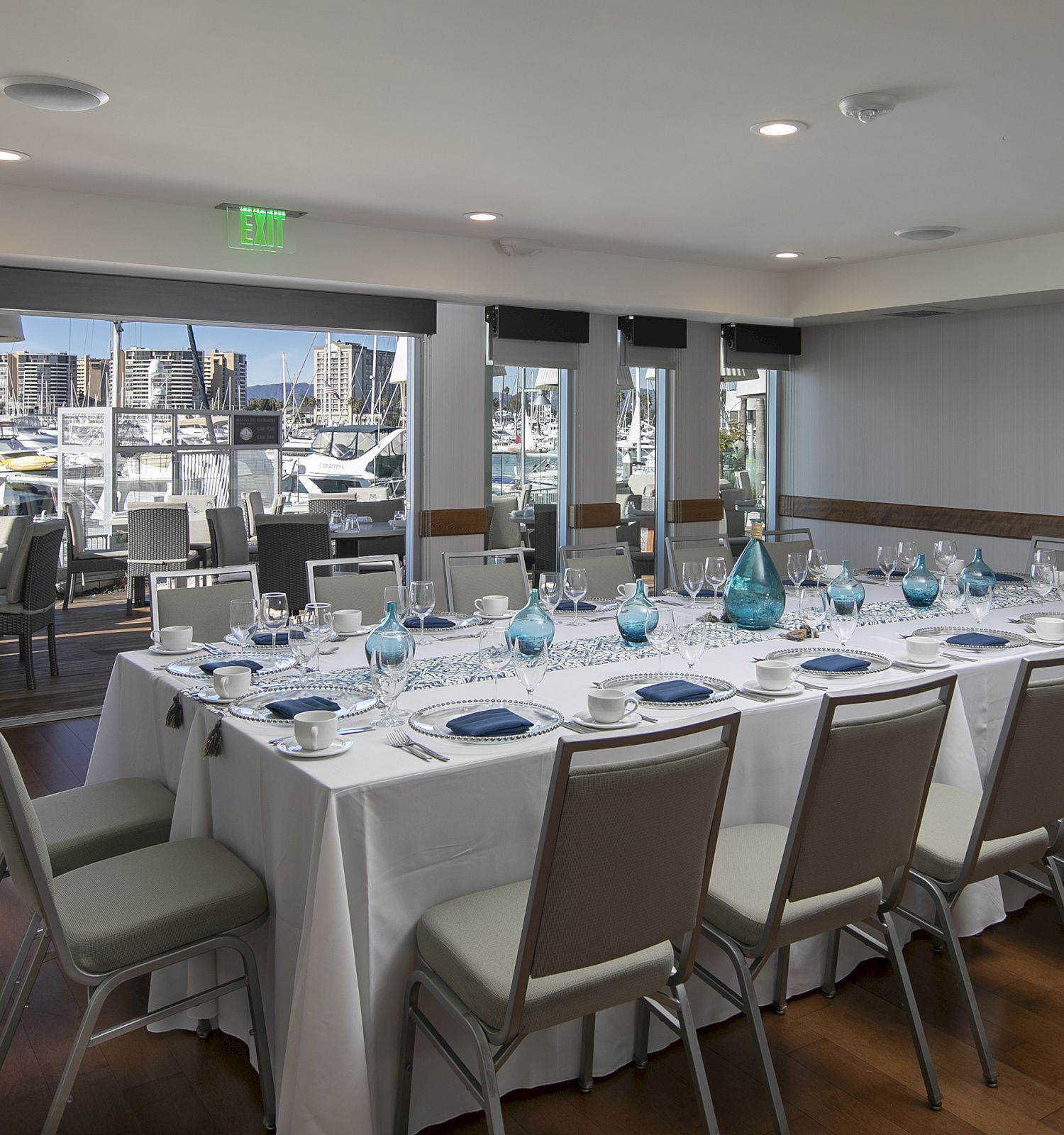 A modern waterfront dining room with a long rectangular table set for a formal meal, white tablecloths, blue glassware, and large windows overlooking boats, ending the sentence.