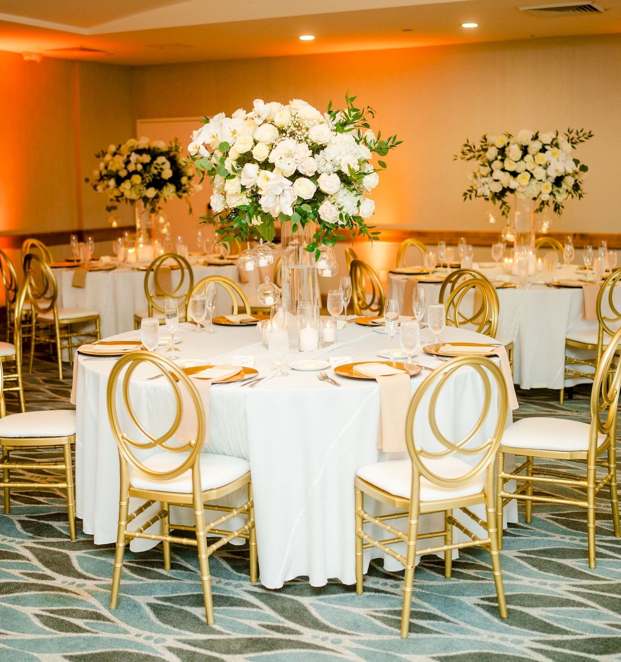 A banquet hall set for a formal event with round tables, white linens, gold chairs, and tall floral centerpieces under warm lighting.