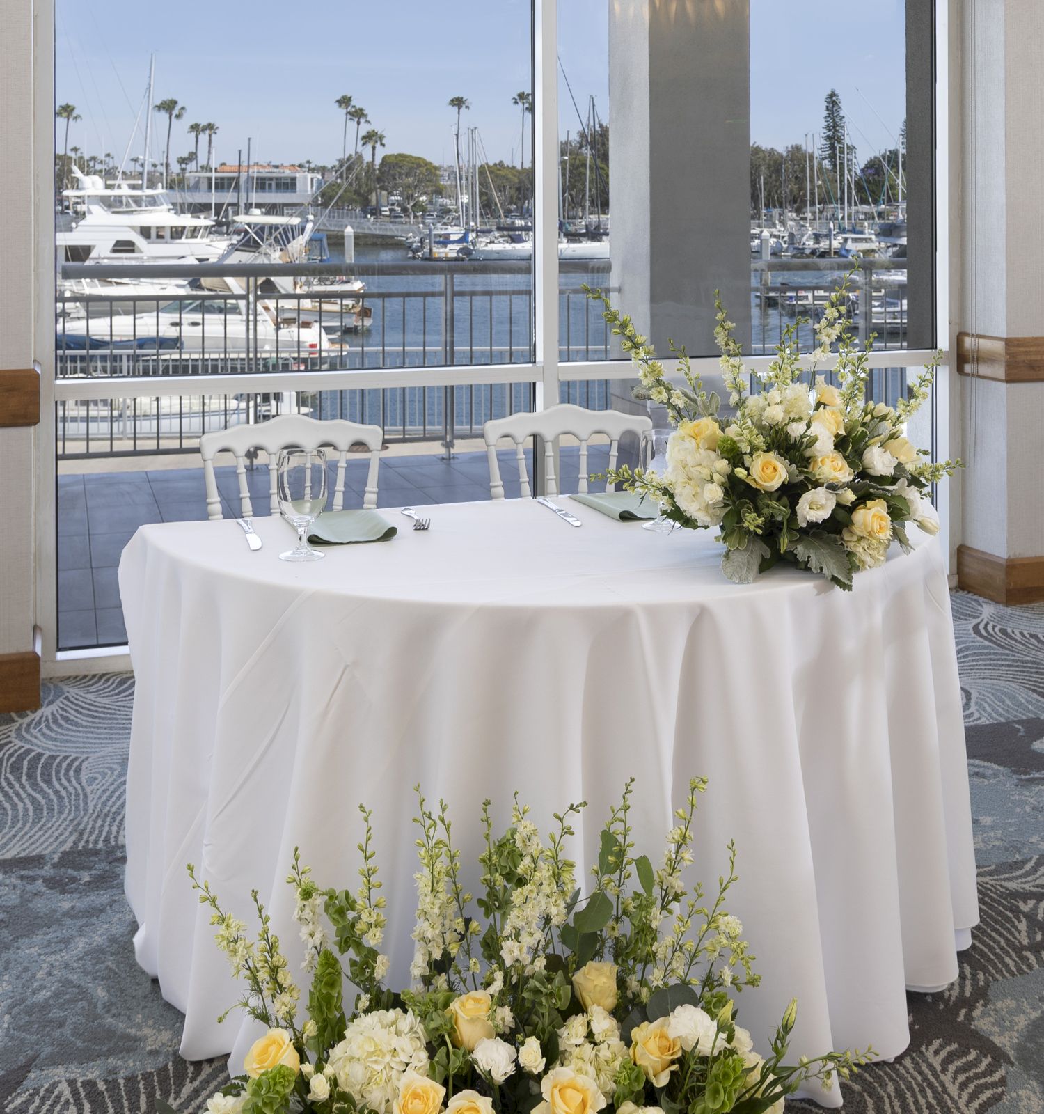 A round table with a white tablecloth, chairs, and a floral centerpiece, set near a window overlooking a marina, for an event.