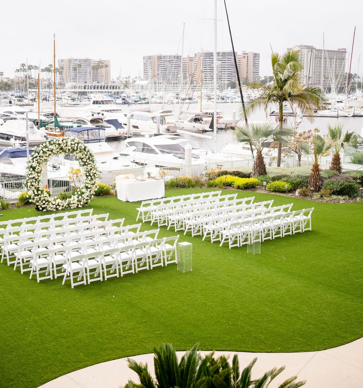 An outdoor wedding setup by the marina: white chairs arranged in rows on green grass, with a curved path, palm trees, and boats in the background.