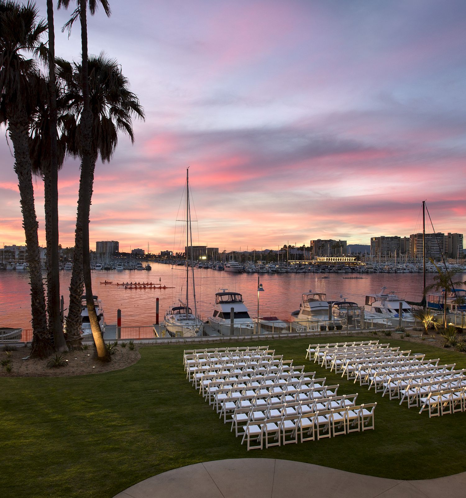 A serene waterfront wedding setup at sunset, with rows of white chairs on a grassy patch facing a calm marina and pink-purple skies.