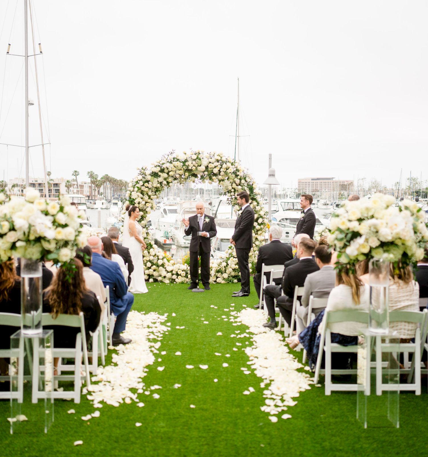 A seaside wedding ceremony with an arch, floral decorations, guests seated on white chairs, and a grassy aisle strewn with petals near boats.