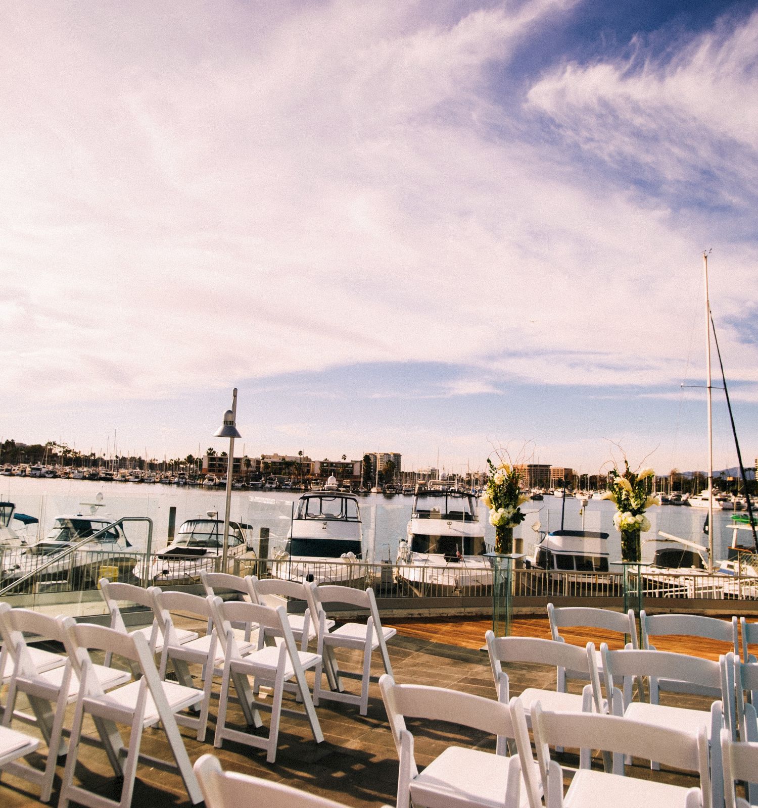 A waterfront event setup with rows of white folding chairs facing a harbor, boats docked along the pier, and a calm sky above.