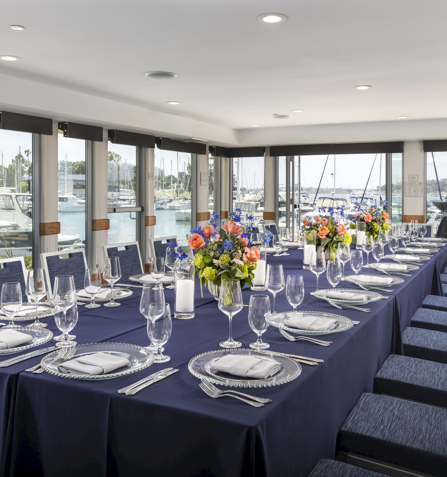 Elegant marina-side dining room with a long, navy-tablecloth setup, place settings, glassware, and colorful floral centerpieces by the windows.