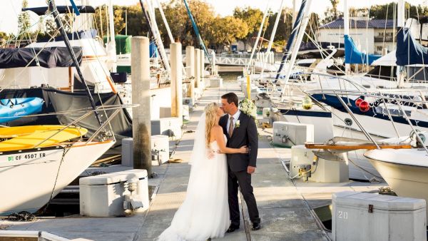 A bride and groom stand on a marina dock, embracing in front of yachts and sailboats as the sun lights the scene.