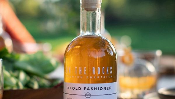 A bottle of The Rocks Old Fashioned cocktail on a table, with glasses and greenery in a sunlit outdoor setting.