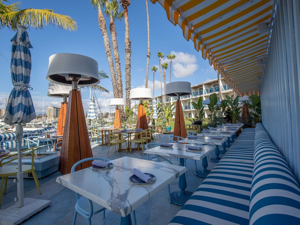 A sunny outdoor dining area at a seaside cafe with blue-and-white stripes, umbrellas, and palm trees by the water.