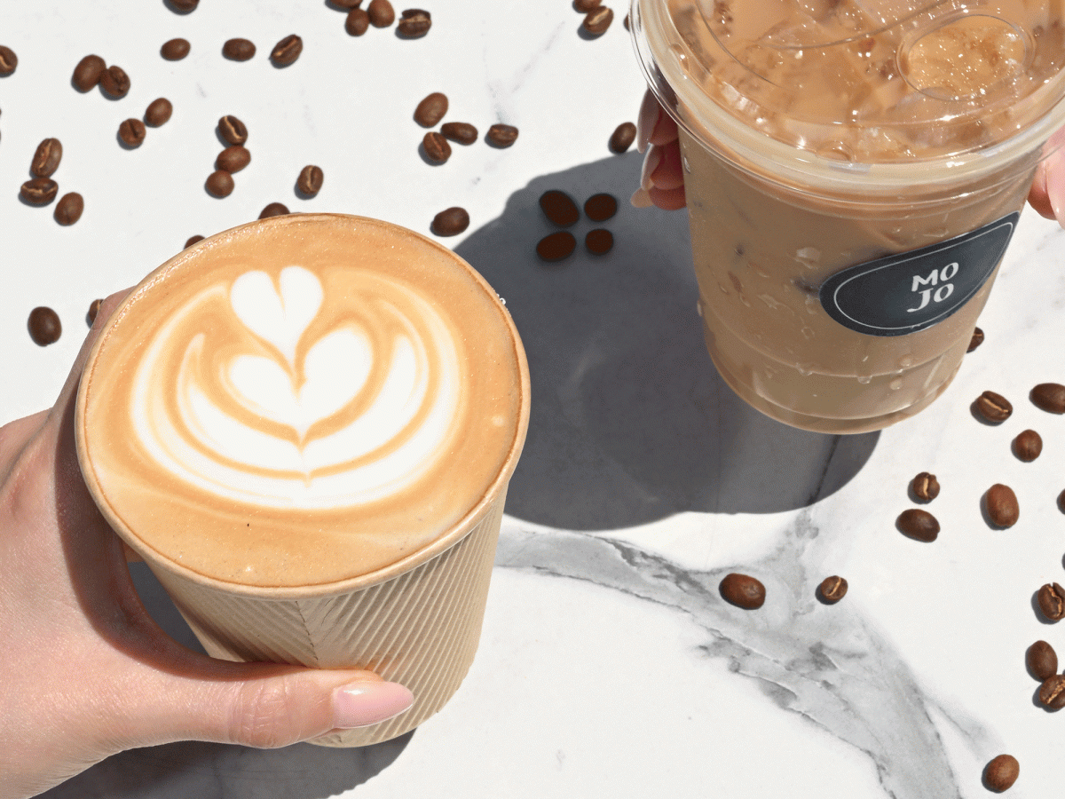 Two iced coffees on a marble surface with scattered coffee beans; latte art crown and foam, shade, and sunlit setup. End with a period.