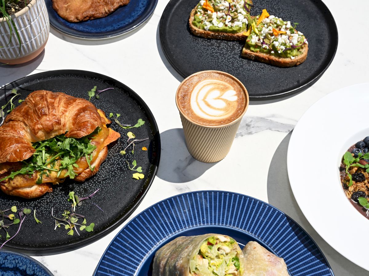 A colorful breakfast spread: croissant sandwich, avocado toast bites, latte art, blueberries, greens, and a plate with grains&mdash;delicious morning feast.