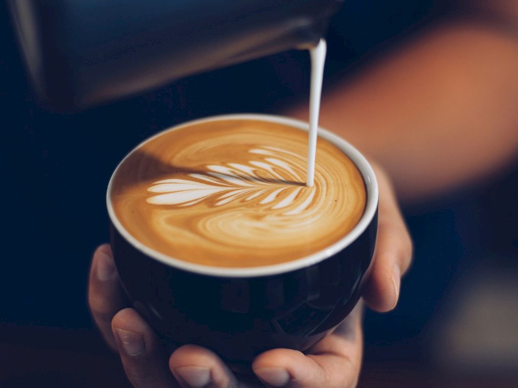 A barista pours steamed milk into a latte with latte art in a dark cup, creating a leaf pattern as the milk blends in.