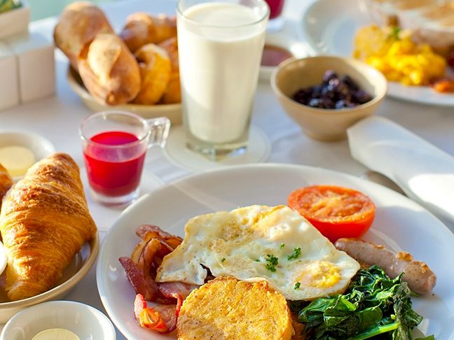 A breakfast spread with eggs Benedict, spinach, toast, croissants, bacon, tomato, hash brown, fruit juice, milk, yogurt, olives, and pastries on a bright table.