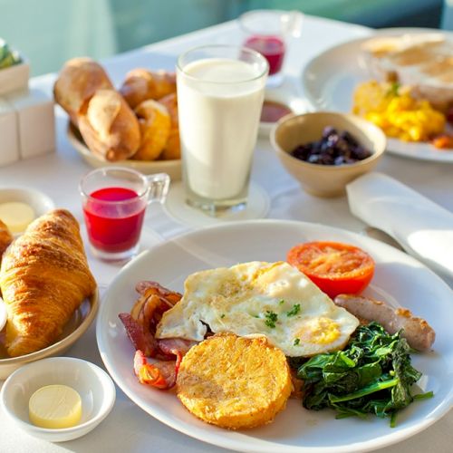 A colorful breakfast spread with toast, fruit, yogurt, a croissant, a pastry, and sunny-side portions on a white table.
