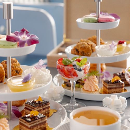 A three-tiered afternoon tea spread with assorted pastries, sandwiches, macarons, fruit, and a cup of tea in the foreground.