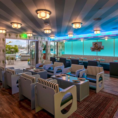 A stylish lounge with striped blue seating, modern tables, and warm lighting; plants along the glass walls create a relaxed, tropical vibe.