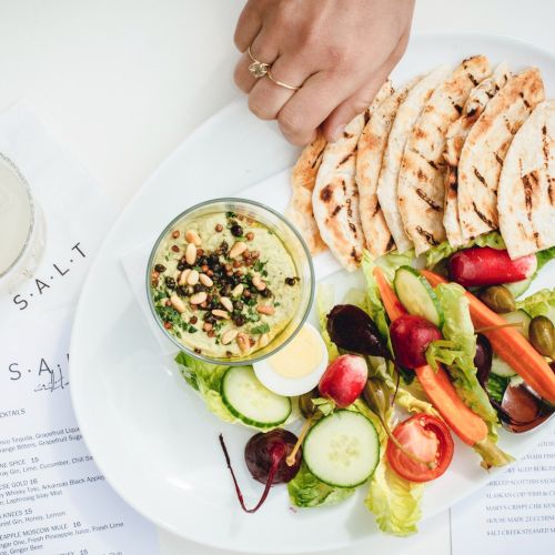 A bright plate with falafel, hummus, cucumber, tomatoes, greens, and pita slices; a lime-garnished drink sits nearby on a white table.