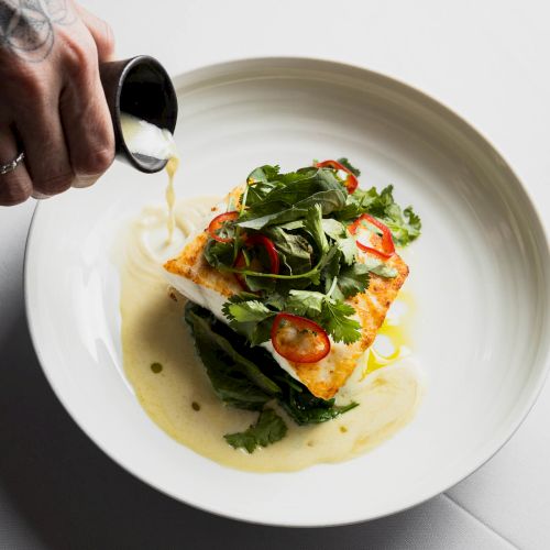 A plated dish with greens on top being drizzled with a light sauce from a small pitcher, highlighting freshness and elegance.