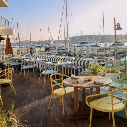 A seaside restaurant patio with striped cushions, wooden deck, yellow chairs, round tables, and a harbour full of sailboats in the background.