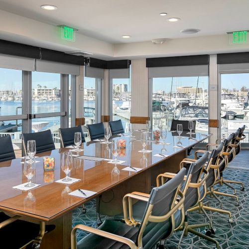 A modern conference room with a long wooden table, laptops, a projector screen, and a few chairs, set for a meeting in a bright, open space.
