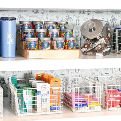 A neat shelf of beverages and snacks: top row shows tall blue bottles, canned drinks, a bag of chips, and a wire basket with boxed items, all organized by type.