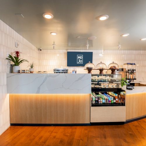 A modern cafe counter with marble top, warm wood panels, and a pastry display; bright white walls, soft lighting, and a polished wooden floor.