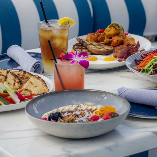 Delicious diners with colorful plates of pasta, a fresh salad, and a bright pink juice, all set on a sunny outdoor table.