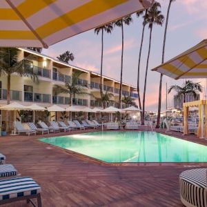 A sunny resort pool area with striped lounge chairs, yellow umbrellas, palm trees, and a modern hotel building in the background, inviting relaxation.