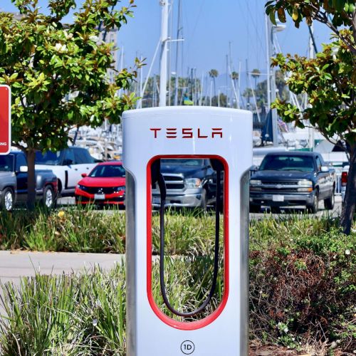 A Tesla Supercharger station with a red-framed charging unit, flanked by two red charging signposts, in a sunny parking lot.