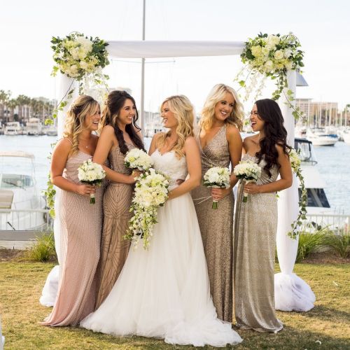 Five women in a wedding scene by the water: a bride in white gown stands with bridesmaids in glittering dresses, holding bouquets under a floral arch.
