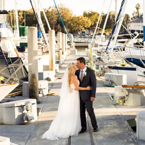 A bride in a white gown and a groom in a suit kiss on a marina dock, surrounded by moored boats and sailboats.